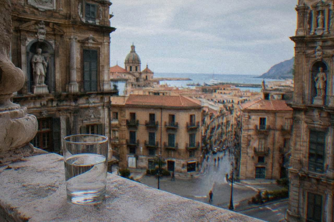 A glass of water standing on a stone ledge overlooking Palermo rooftops and the sea