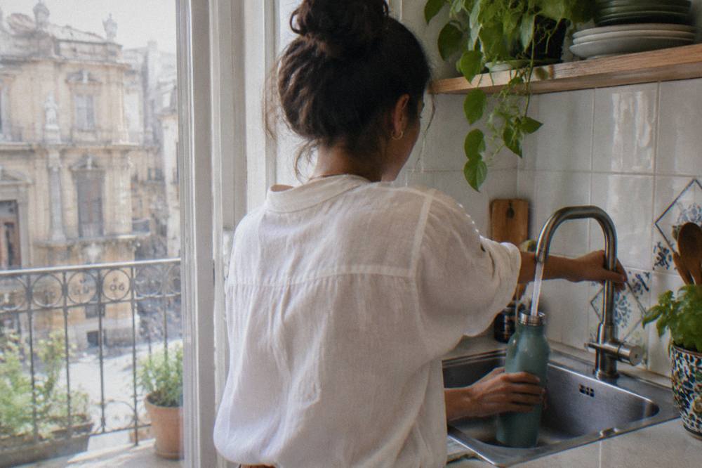 A person filling a reusable water bottle from a kitchen tap in a bright Palermo apartment