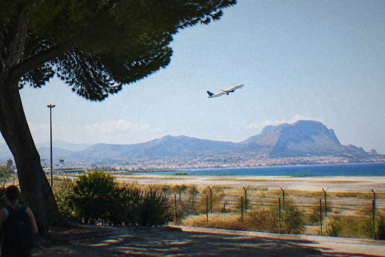 An airliner climbing over a coastal Sicilian city with mountains behind the bay