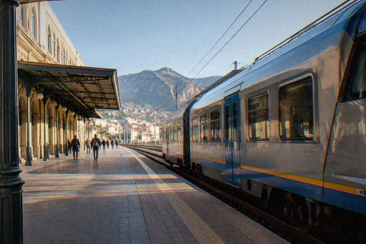 A regional train waiting at a Sicilian railway station platform under a clear sky