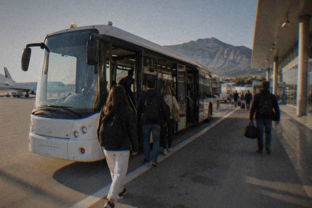 Passengers boarding a white airport shuttle bus at the curb outside an airport terminal in Sicily