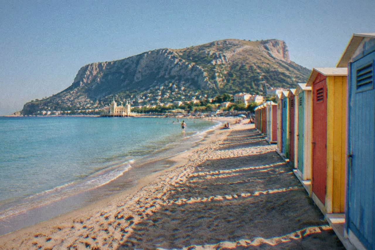 A wide sandy beach with calm turquoise water and a row of colourful changing cabins along the shore