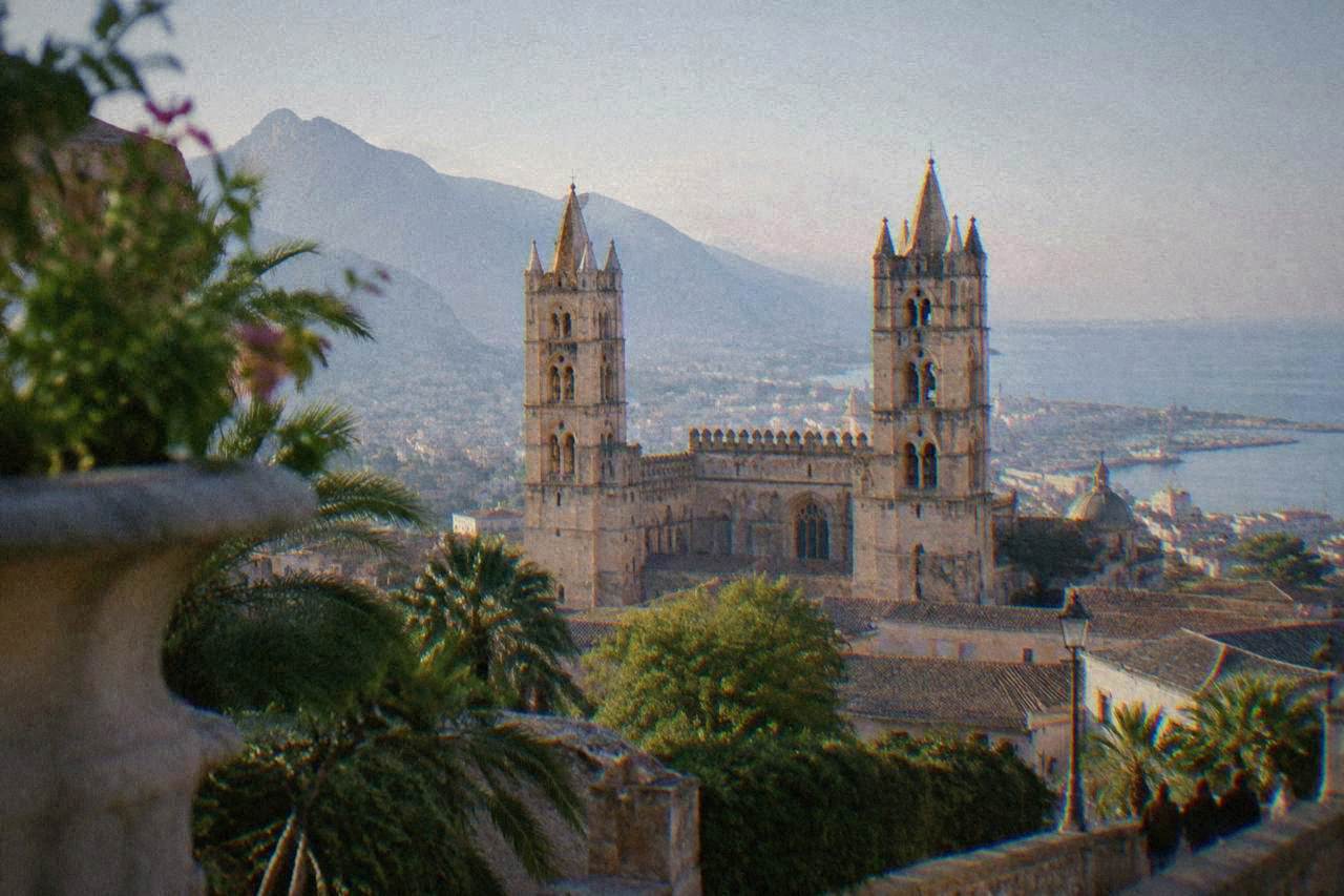 A hilltown cathedral with twin towers rising above rooftops on a mountainside above a coastal plain