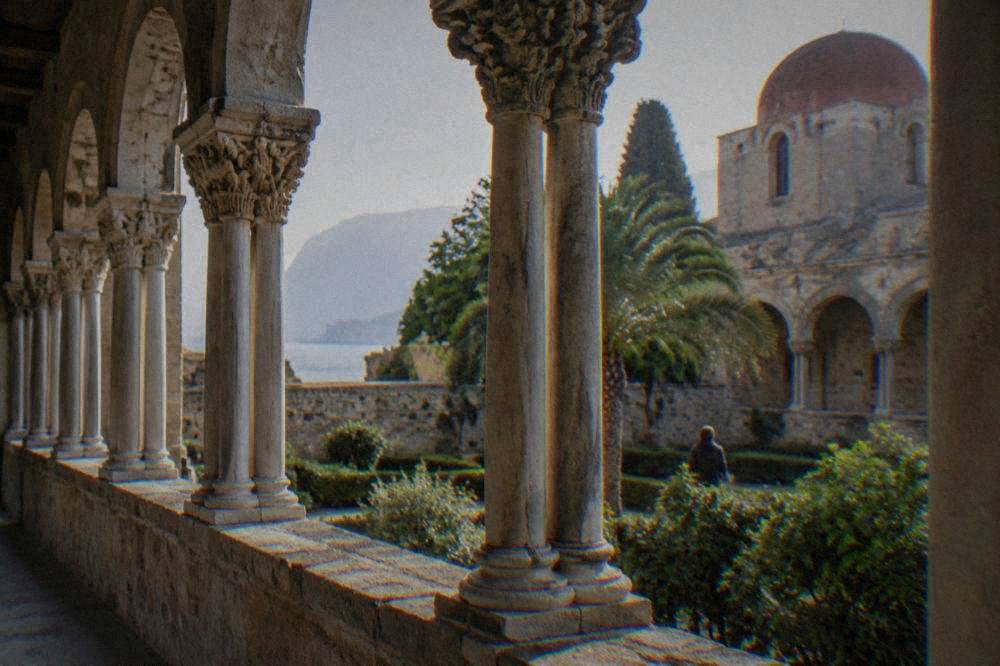 A stone cloister with paired columns and carved capitals surrounding a garden courtyard