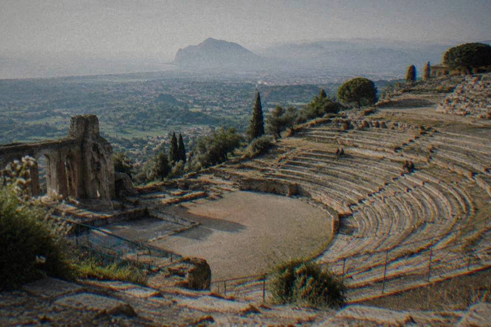 A stone theatre carved into a hillside overlooking a wide green valley in Sicily