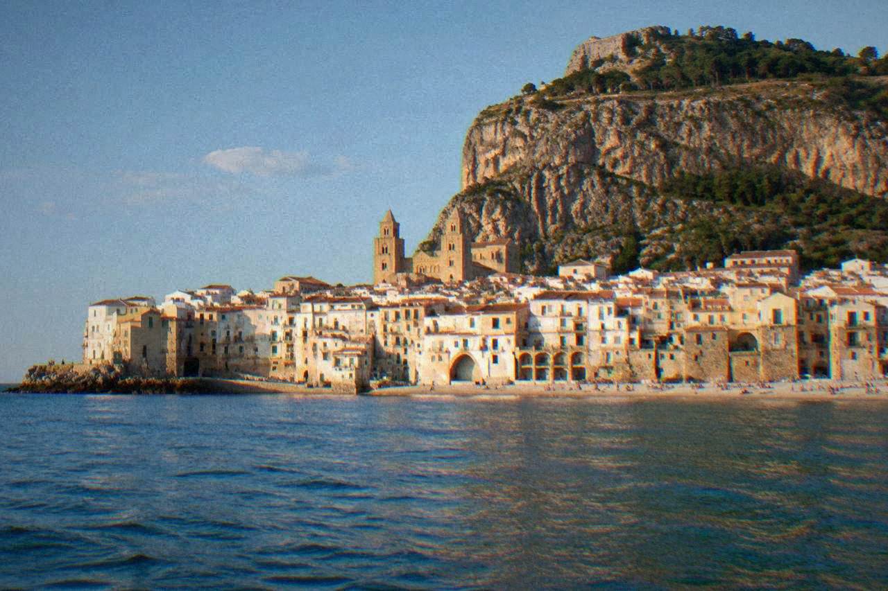 The sandy beach and packed houses of Cefalu seen from the sea, with the rocky headland behind the town