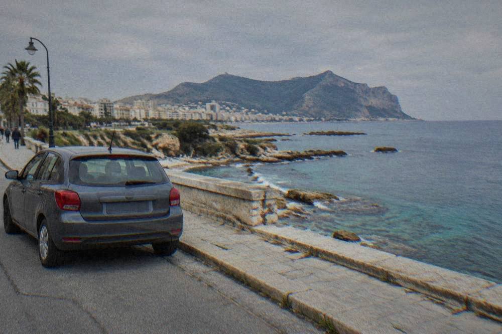 A car parked on a seafront road above a rocky Sicilian coastline with clear water