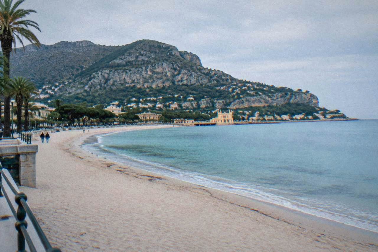A wide sandy beach curving around a calm blue bay with a promenade and wooded hills behind it