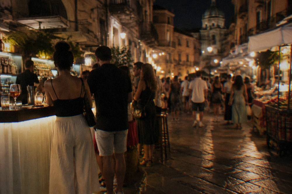 Lit bar counters and people standing with drinks in a Palermo market street at night