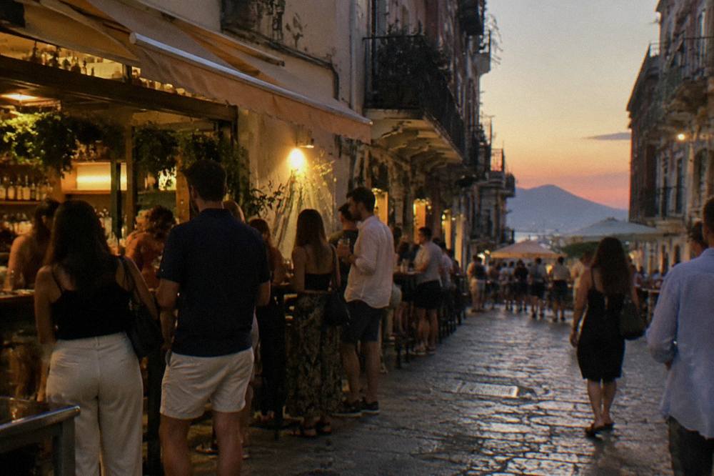 An evening street in Palermo with lit bar fronts and people standing with drinks