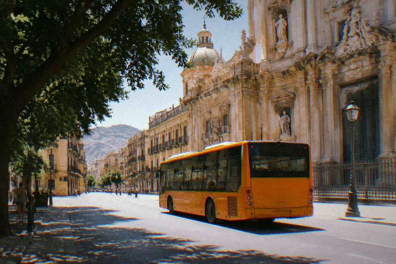 An orange AMAT bus passing baroque buildings on a sunlit street in central Palermo