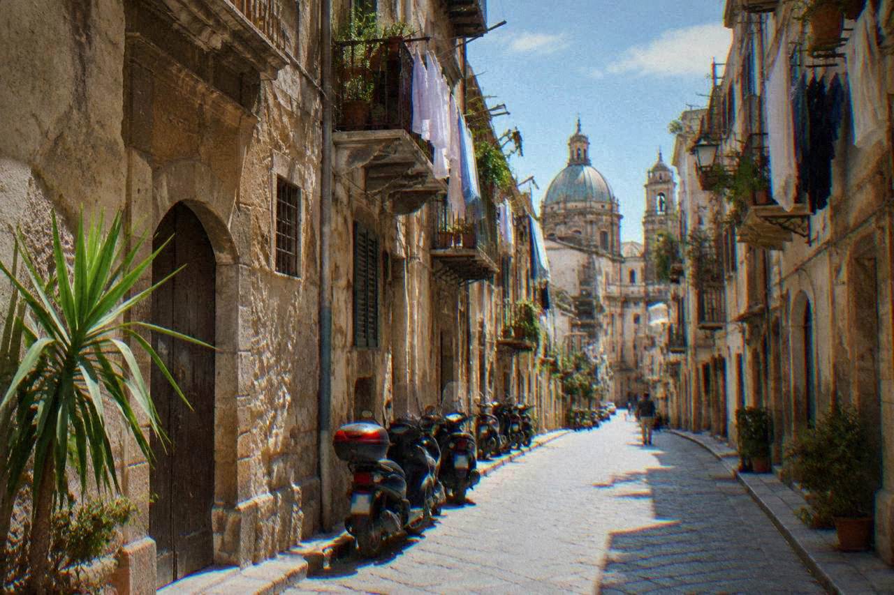 A narrow Palermo street with worn stone buildings, scooters parked against the kerb and laundry hung from balconies