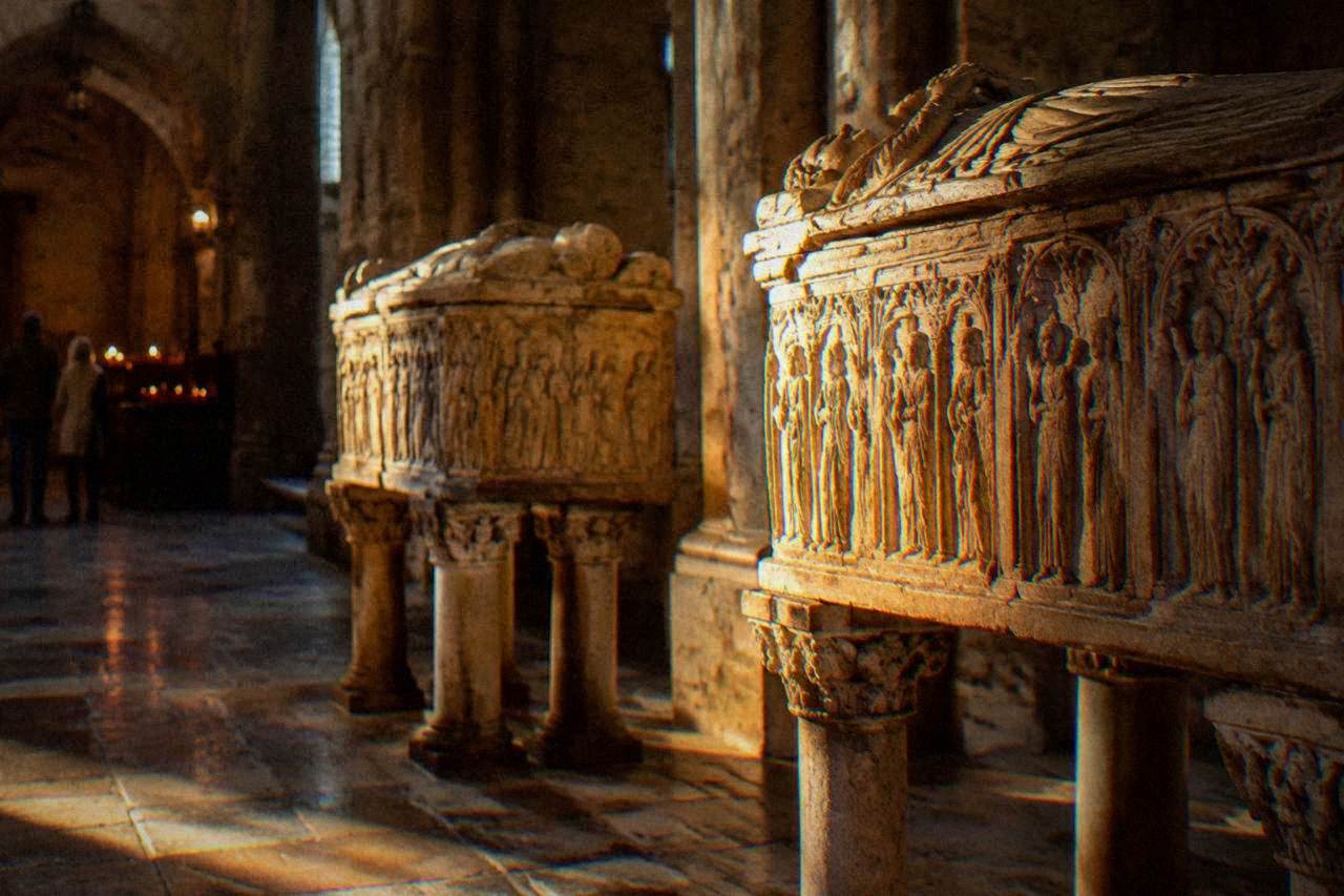 Carved stone sarcophagi resting on columns inside a dim cathedral chapel