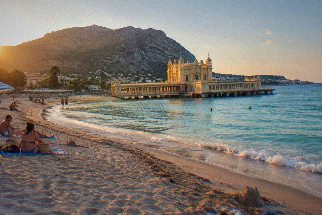 A wide sandy beach curving around a turquoise bay with a historic bathing pavilion at its centre