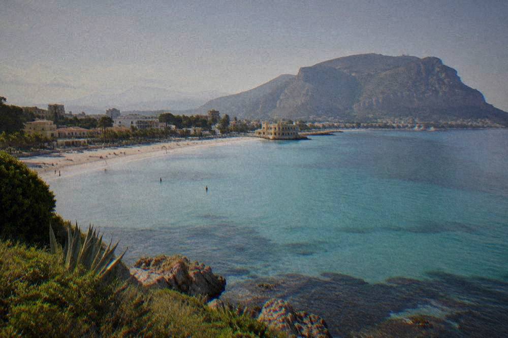 A sandy bay seen from a headland with turquoise shallows and a town behind the beach