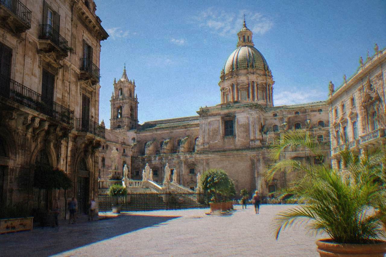 A sunlit square in the historic centre of Palermo with ornate old buildings and a church dome