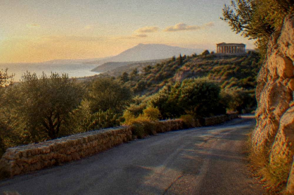 A country road winding through Sicilian hills toward distant Greek temple columns