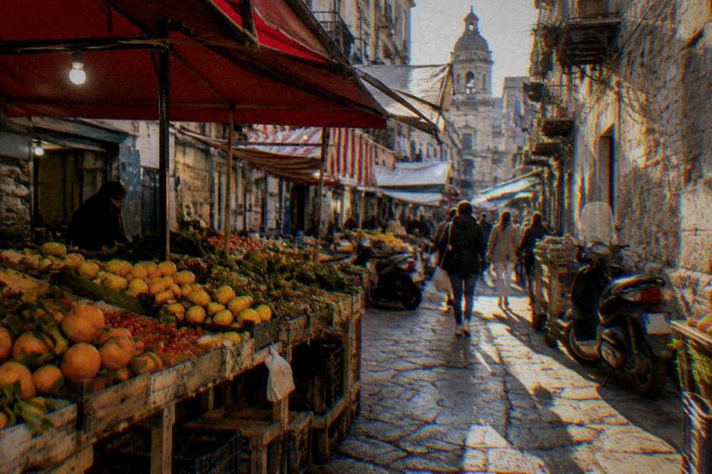 A narrow Palermo street with market awnings, scooters and shoppers in the Albergheria district