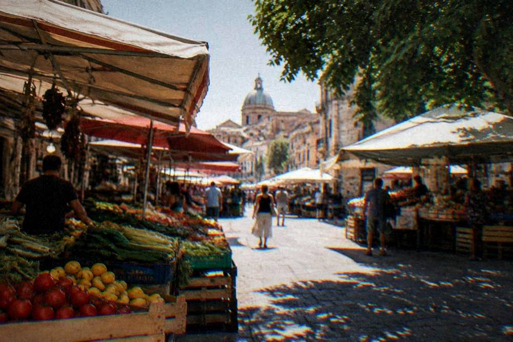 Shoppers and vendors under market awnings in a sunlit Palermo street in the late afternoon