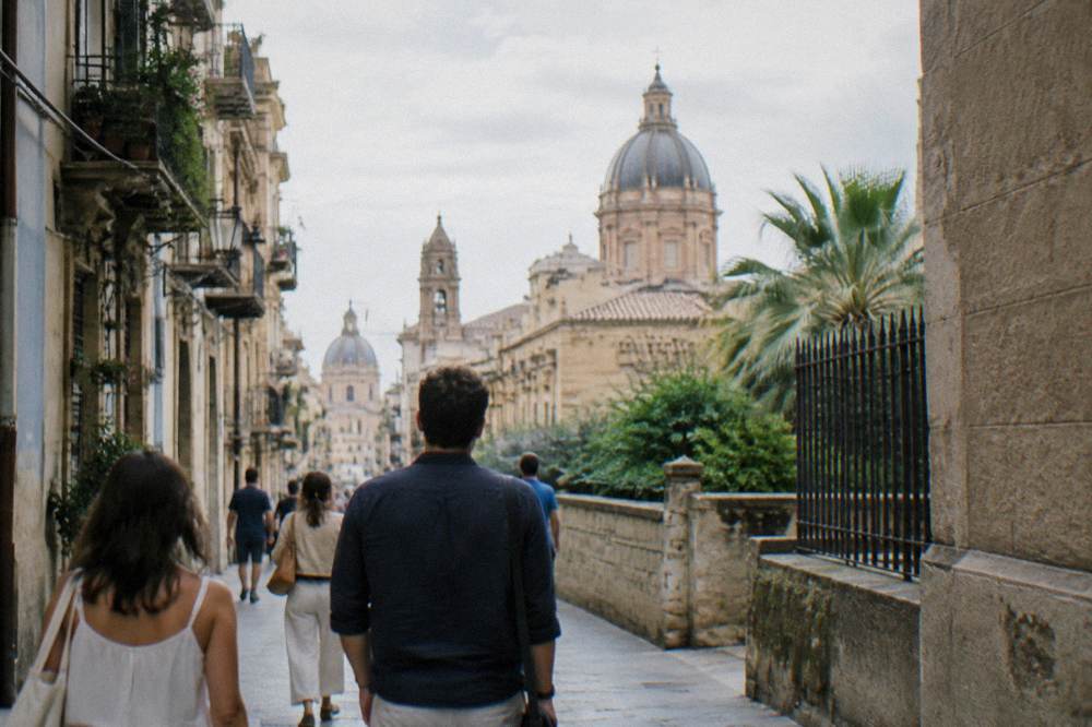 People walking along a sunlit Palermo street with church domes on the skyline