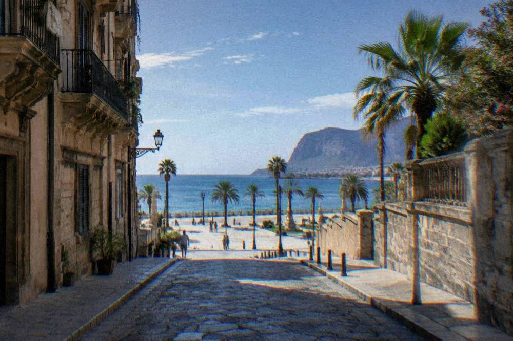A flat historic street in Palermo leading downhill toward a bright open seafront square