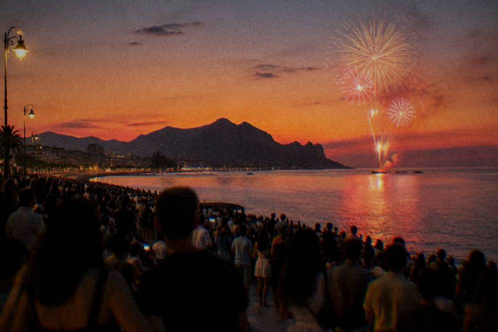 A large evening crowd gathered on a seafront esplanade with fireworks bursting over dark water