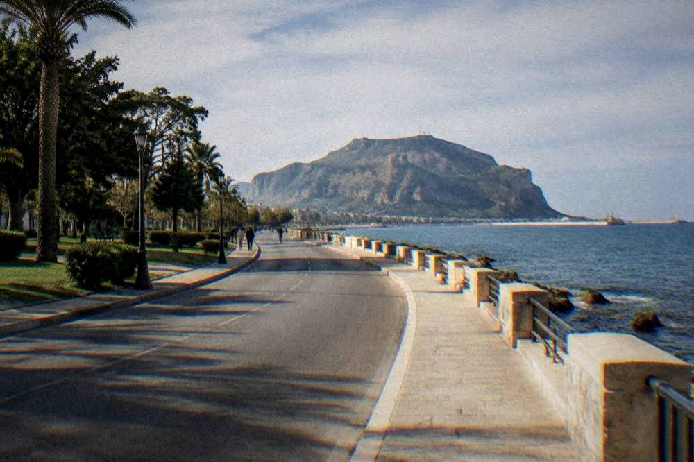 A wide coastal road curving between a green park and the open sea toward a distant mountain