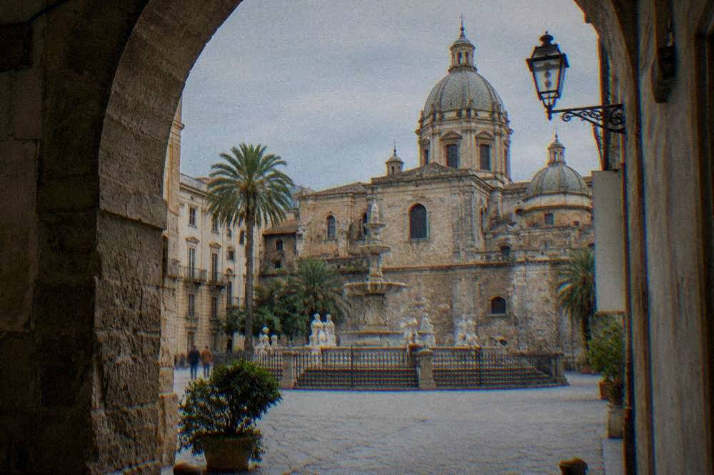 Sunlit square with a fountain and church domes seen through an archway
