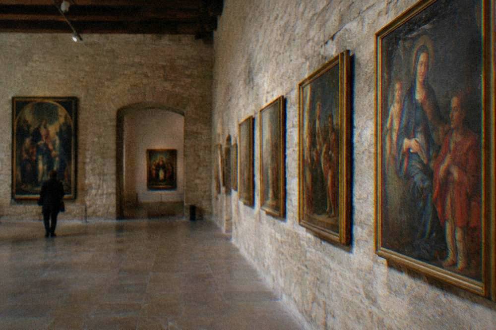 A gallery room with stone walls and framed paintings hung in rows