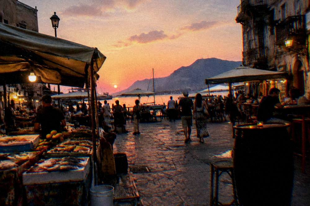An evening street market scene with food stalls and lit bars in an old Palermo square