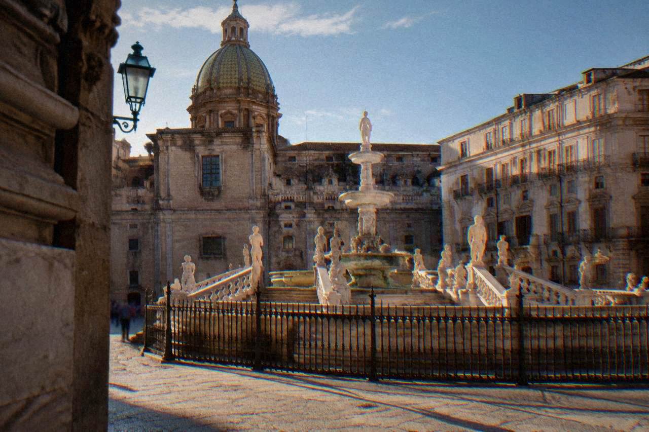 A grand tiered stone fountain with statues standing in a sunny square lined with church facades and historic buildings