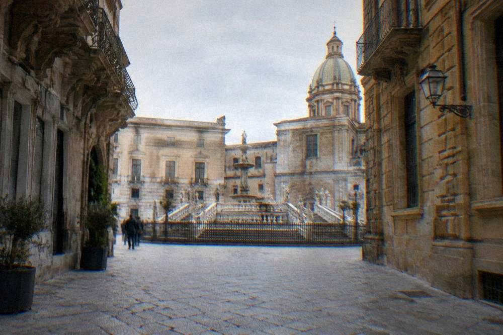 A pedestrian street in an old Sicilian town with ornate buildings on both sides leading toward a fountain square