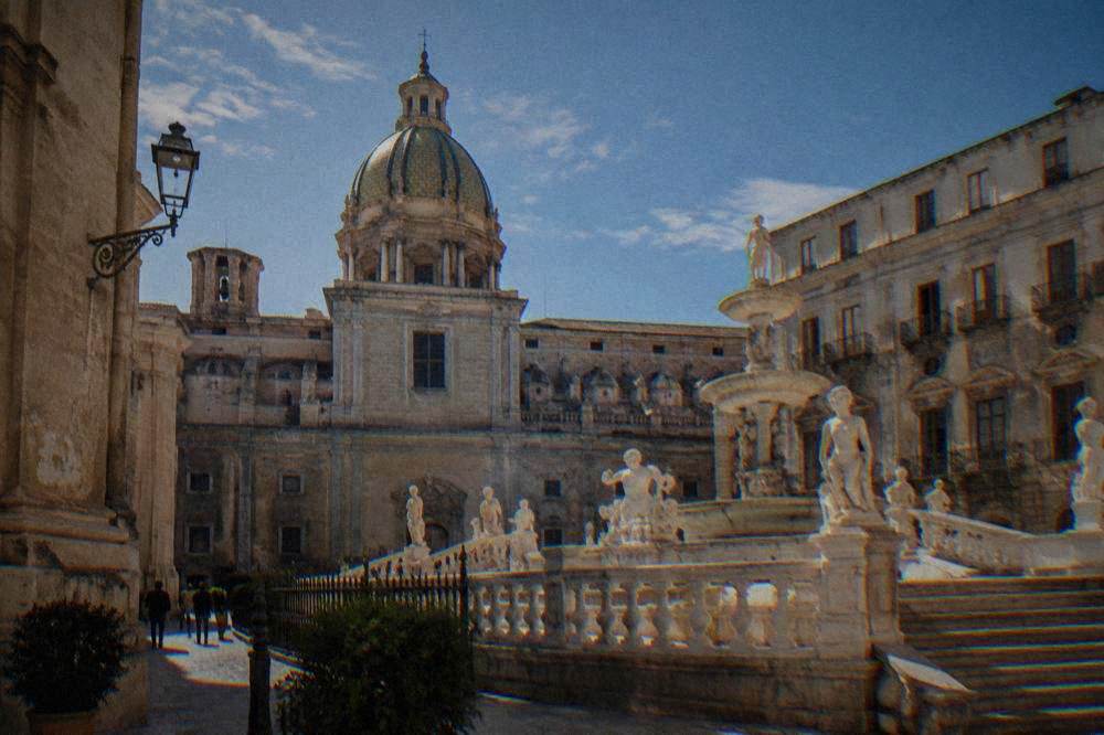 Ornate church facade and historic palazzo walls framing a fountain square in a Sicilian city