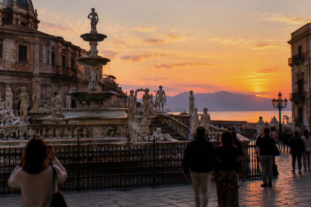 People strolling and photographing a large ornate fountain in an evening city square