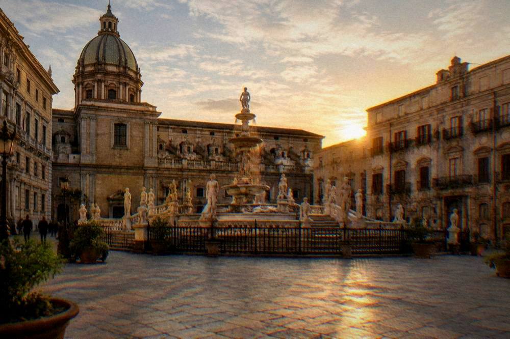 Wide view of a Sicilian city square at golden hour with an ornate fountain and historic facades