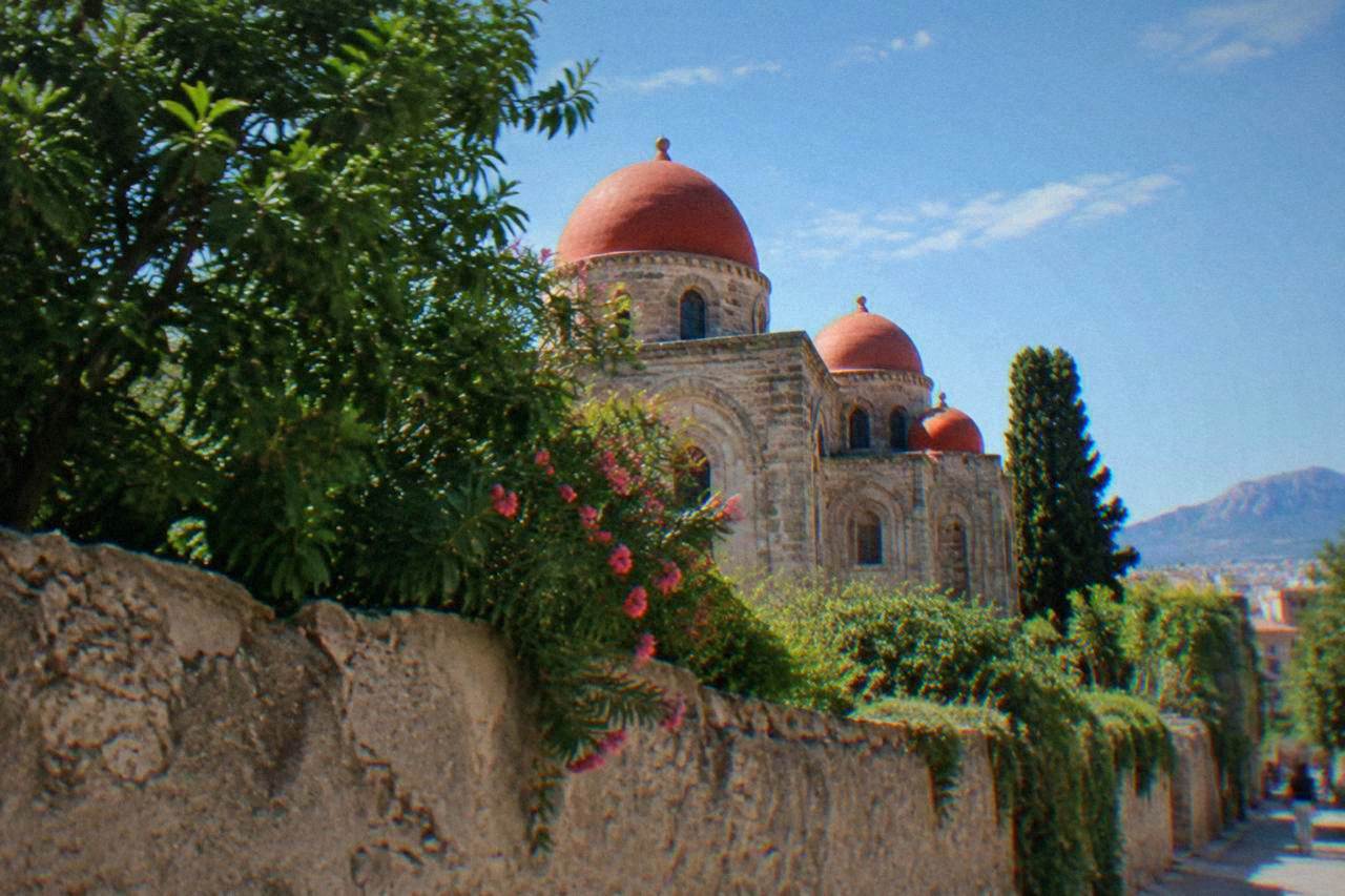 A small church with red spherical domes seen over a garden wall in central Palermo