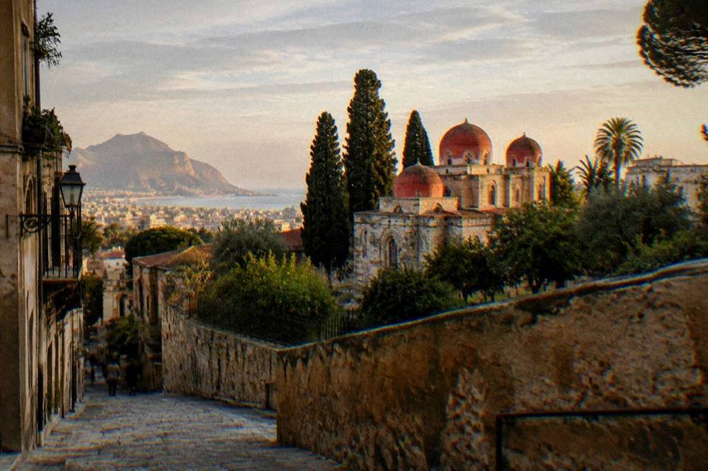 The walled garden and red domes of a small church seen from a sloping street in Palermo at golden hour