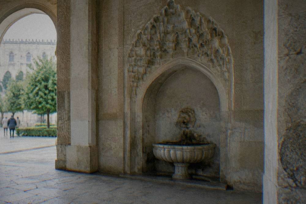 An arched niche with a wall fountain inside a stone hall, muqarnas decoration above the arch