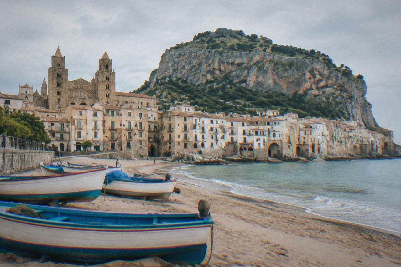 Fishing boats resting on the sandy beach below the old town of Cefalu with the rock of La Rocca rising behind the buildings