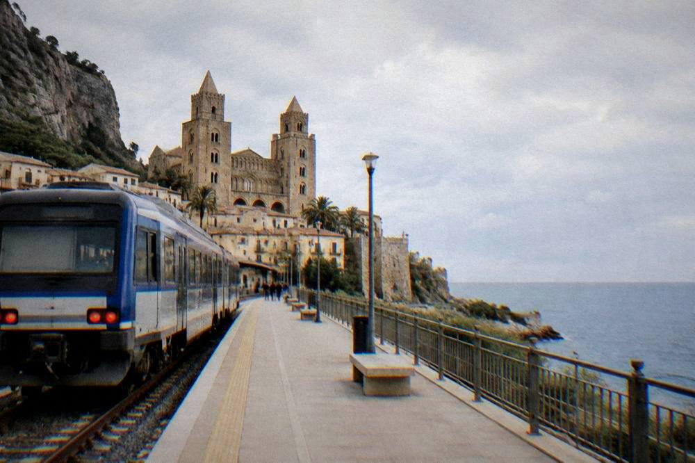 A regional train pulling away from a small coastal station with the sea visible beyond the platform