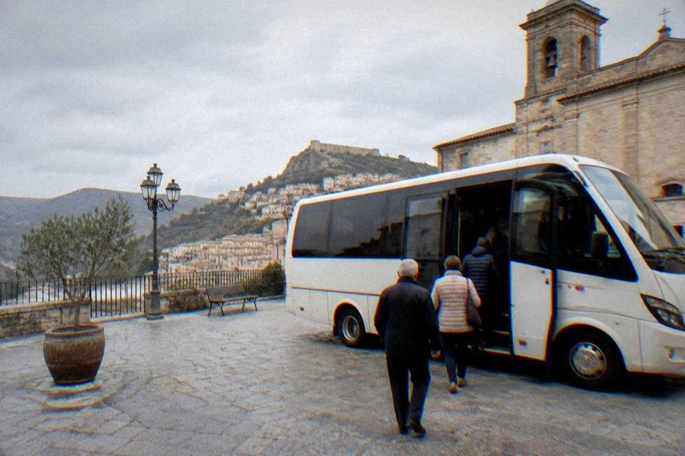 A small coach parked on a stone-paved square in a Sicilian hill town with passengers boarding
