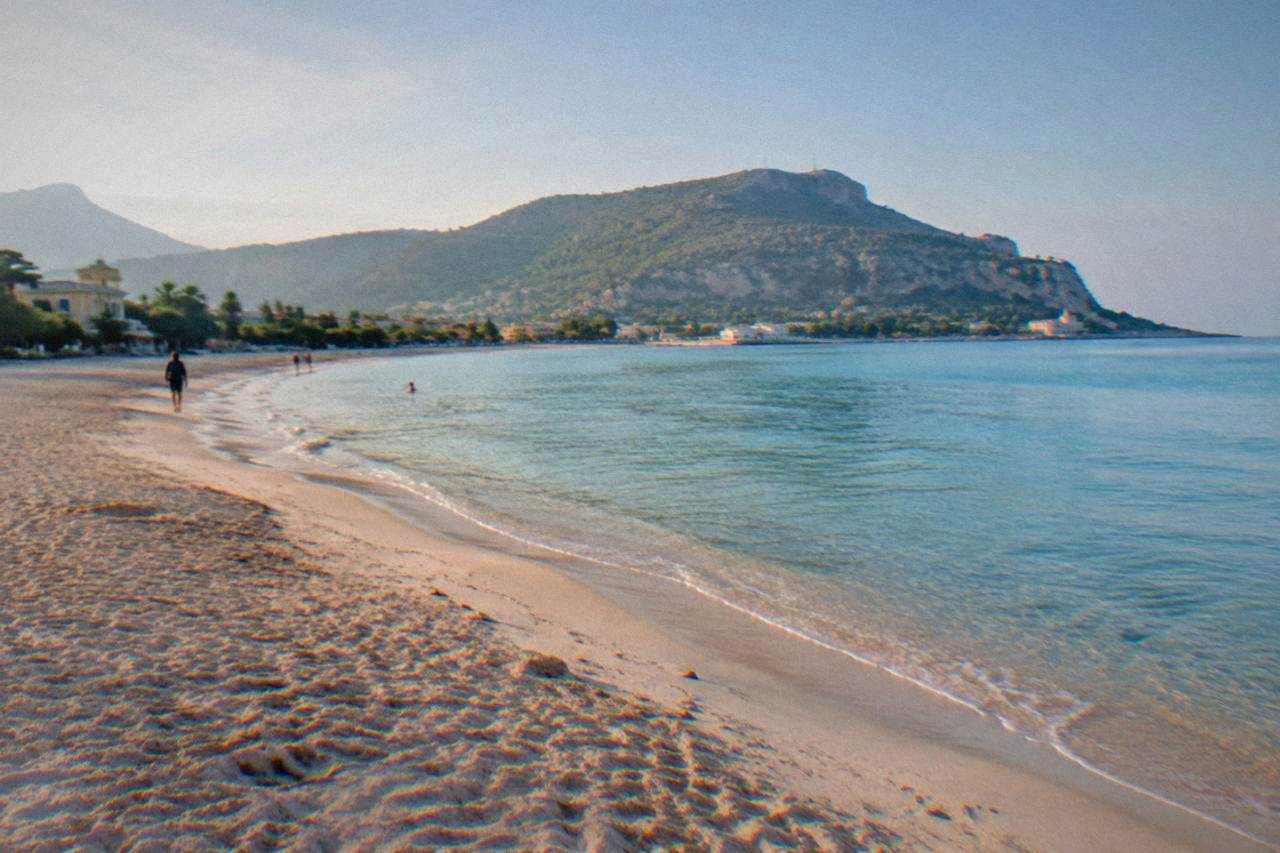 A wide sandy beach curving around a calm blue bay with a wooded headland in the background