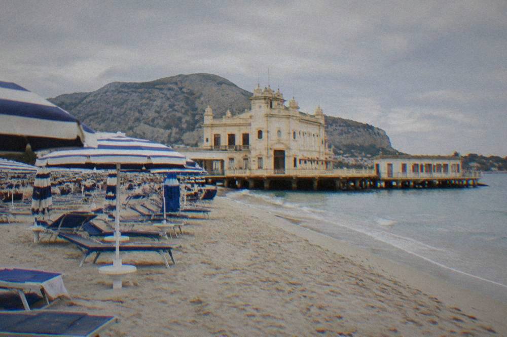 Rows of sun loungers and striped umbrellas on sand in front of a historic bathing pavilion