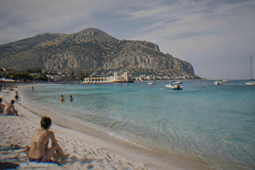 People on white sand beside turquoise shallows with small boats moored in the bay