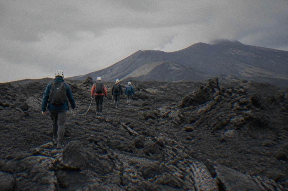A small roped group of hikers with helmets walking across a grey lava field on Etna
