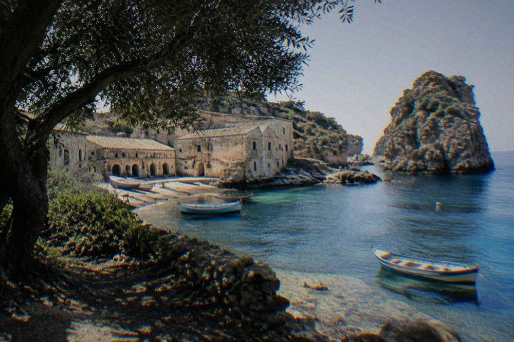 Old stone buildings of a former tuna fishery beside a clear blue cove with wooden boats