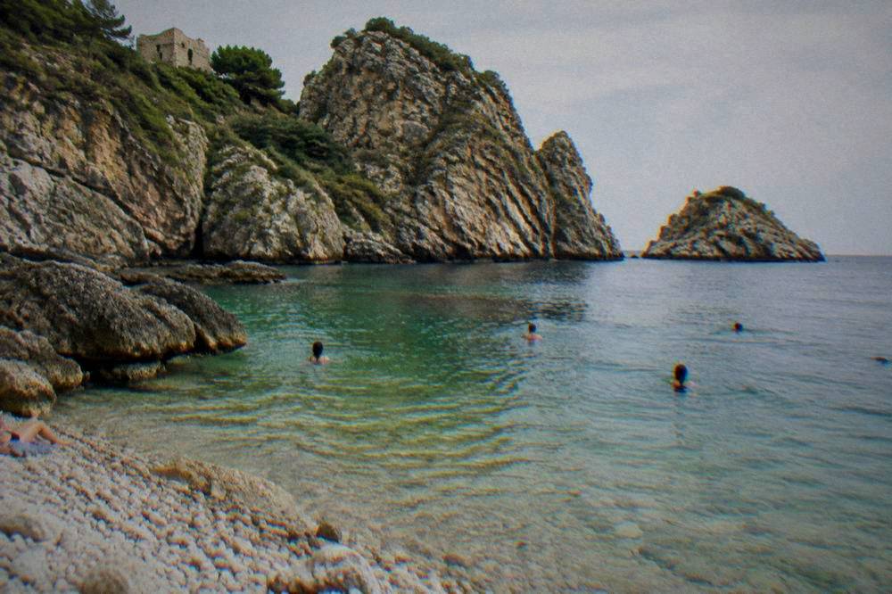 People swimming from a pebble cove in clear green water with rocky cliffs behind