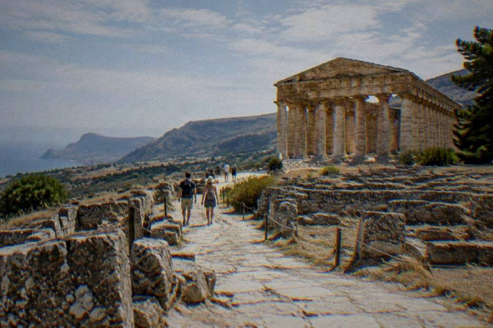 Visitors walking a stone path among ancient ruins under strong summer sun