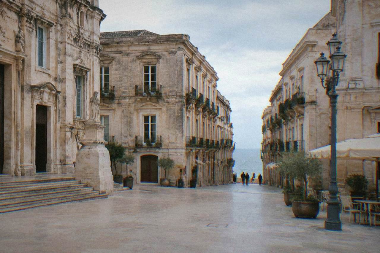 View across a sunlit square in Ortigia with old stone buildings and a narrow street leading to the sea