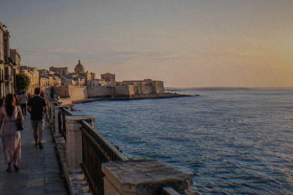 People walking along a sea wall promenade beside deep blue water at golden hour